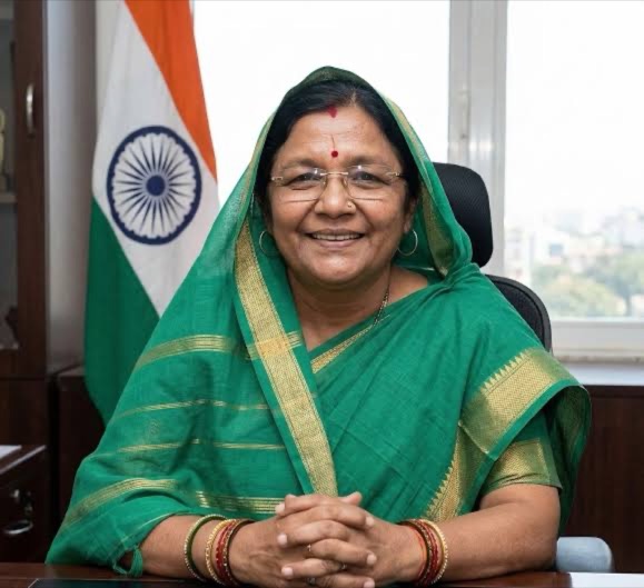 Smt. Savitri Thakur seated at her office desk in a green sari, with the National Flag of India behind her.