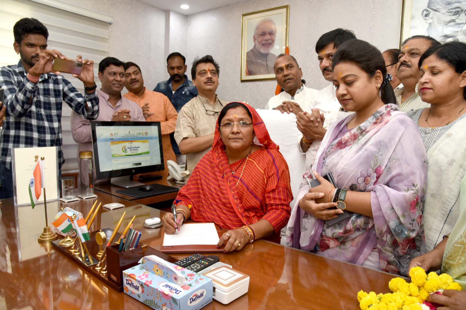 Smt. Savitri Thakur signing documents at the ministerial desk as she assumes charge, with officials and colleagues present.