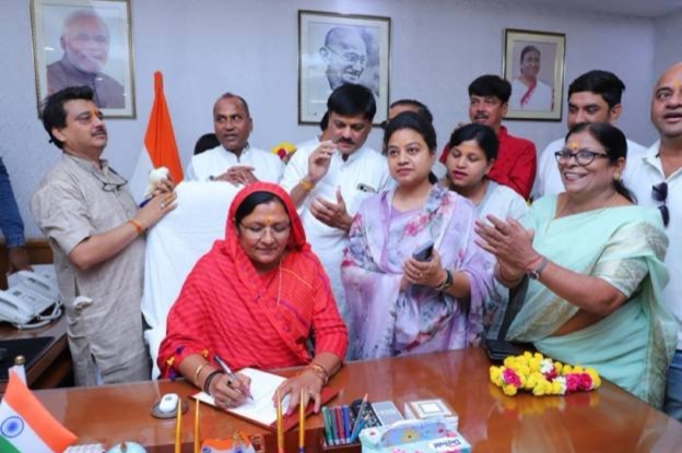 A wider view of the ministerial office as Smt. Savitri Thakur signs papers, with framed portraits on the wall behind.