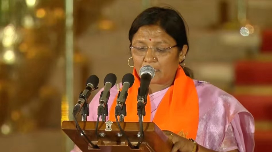 Smt. Savitri Thakur reading the oath of office at a lectern bearing the State Emblem of India.