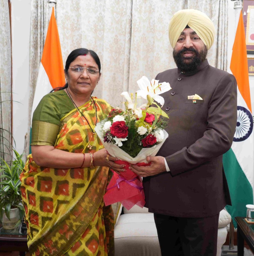 Smt. Savitri Thakur receiving a bouquet from a State Governor in an official reception room, flanked by the National Flag.