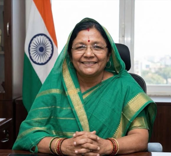Smt. Savitri Thakur seated at her office desk in a green sari, with the National Flag of India behind her.