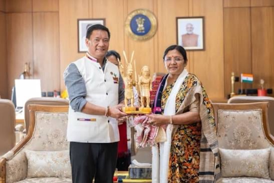 Smt. Savitri Thakur receiving a carved wooden memento from a State Chief Minister in an official reception room.
