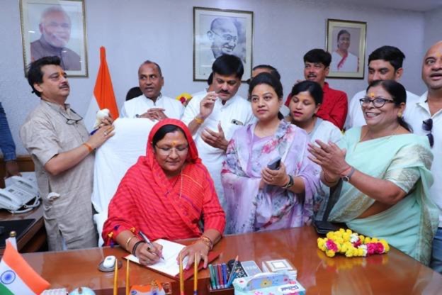 Smt. Savitri Thakur signing a document at her ministerial desk, surrounded by officials and colleagues, as she assumes charge.